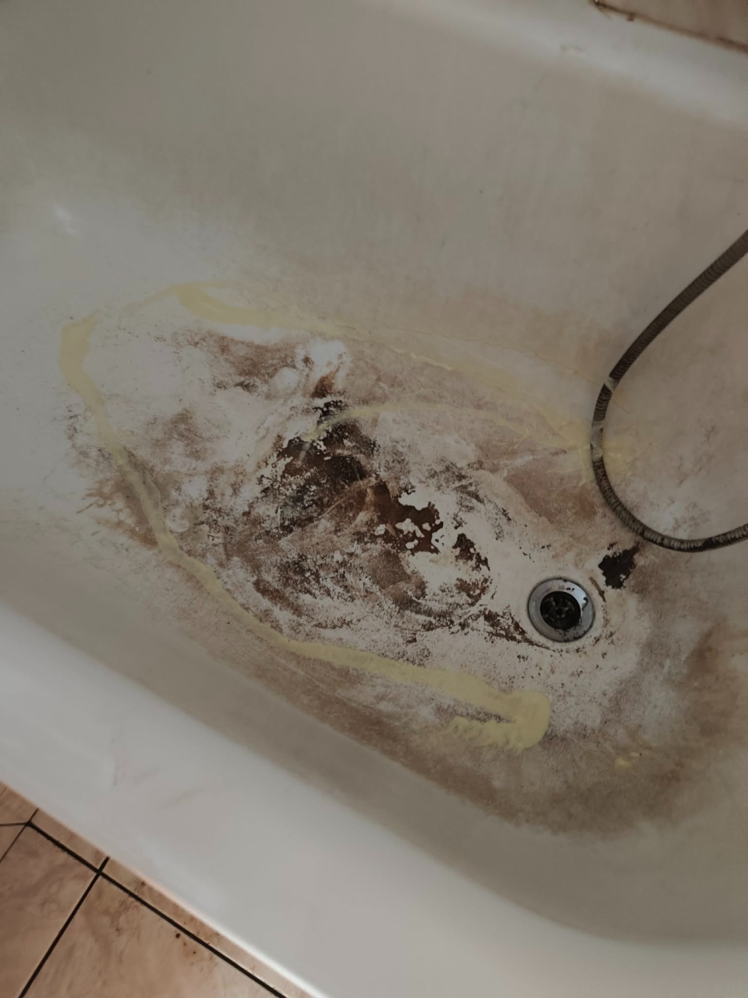 Zaniedbana wanna z widocznymi zabrudzeniami, osadami i plamami. W odpływie ciemne zanieczyszczenia. Wąż prysznicowy w prawym górnym rogu. Brudna wanna przed czyszczeniem.