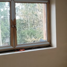 Tynki maszynowe Kamil Ziobro - Okna z widokiem na zieleń po tynkowaniu, widoczne ślady prac na parapecie i ścianie. Nowe tynki w jasnym odcieniu, gniazdko elektryczne na dole ściany.