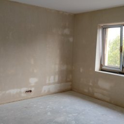 Tynki maszynowe Kamil Ziobro - Pokój po tynkowaniu maszynowym, widoczne gładkie ściany w odcieniach beżu, okno z widokiem na zieleń i gniazdka elektryczne. Świeżo położona podłoga.