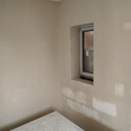 Tynki maszynowe Kamil Ziobro - Świeżo otynkowane wnętrze z oknem, widoczne ślady po łatach. Na pierwszym planie fragment stołu roboczego. Neutralna kolorystyka ścian i sufitu.