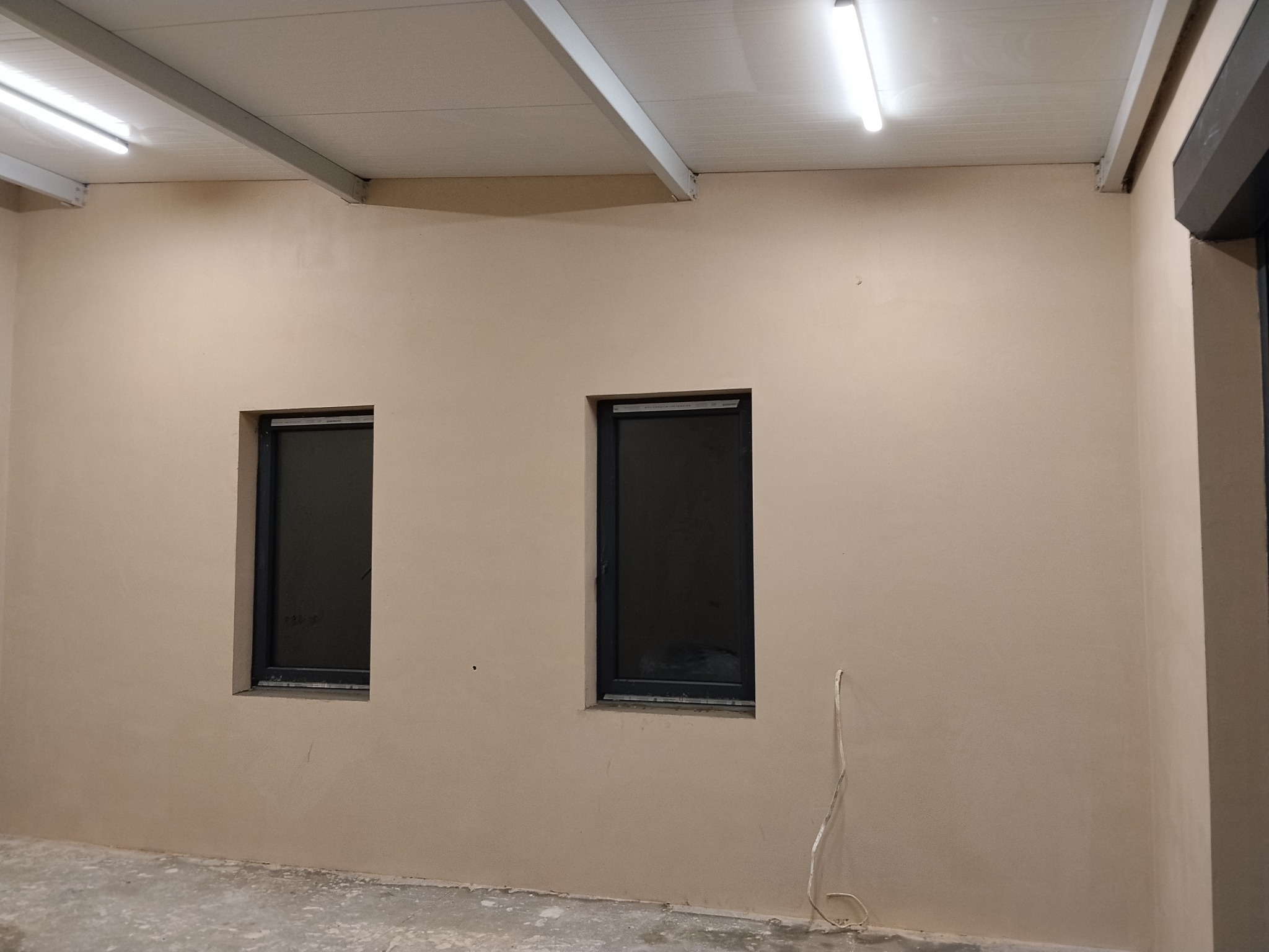 Wnętrze po nałożeniu gładzi: ściany w beżowym odcieniu z dwoma oknami w czarnych ramach. Widoczne ślady po pracach wykończeniowych na podłodze i wystający kabel.