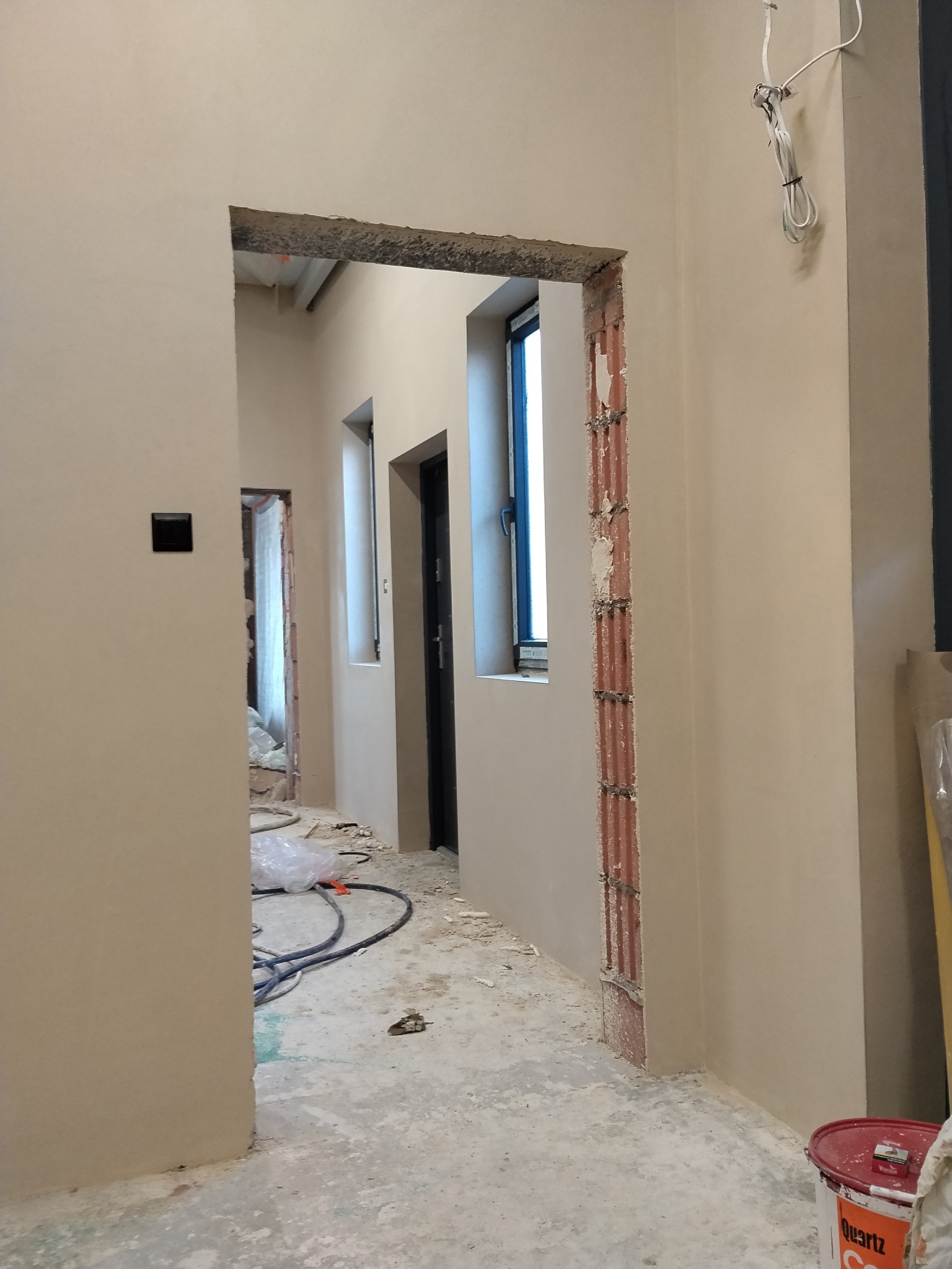 Wnętrze po nałożeniu jasnej gładzi, widoczne surowe ościeżnice okien i drzwi, kabel zwisający ze ściany, podłoga z resztkami gruzu i wiadro farby.