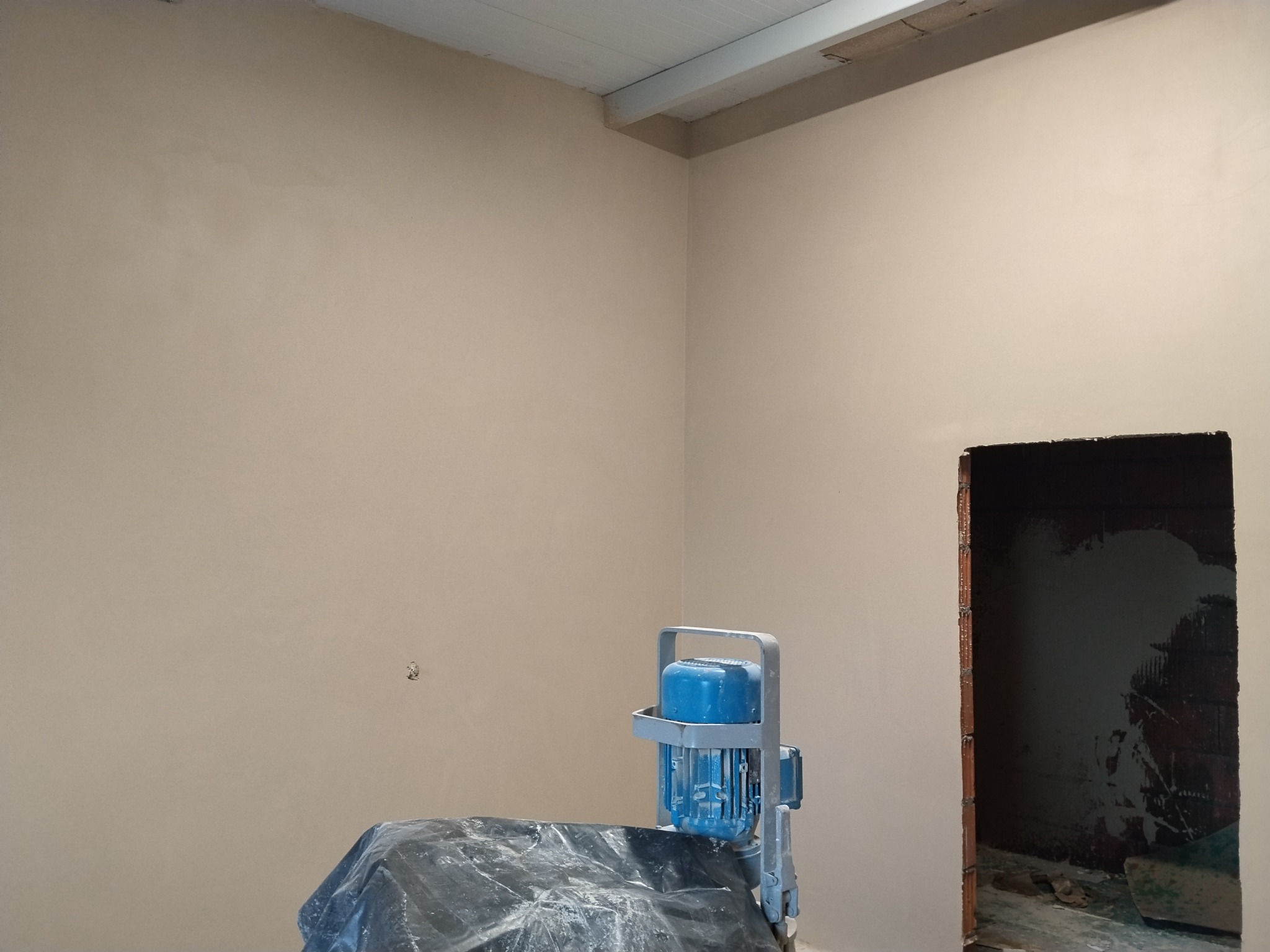 Wygładzone ściany w rogu pomieszczenia, gotowe do malowania. Na pierwszym planie maszyna przykryta folią. Widoczny otwór drzwiowy w stanie surowym.