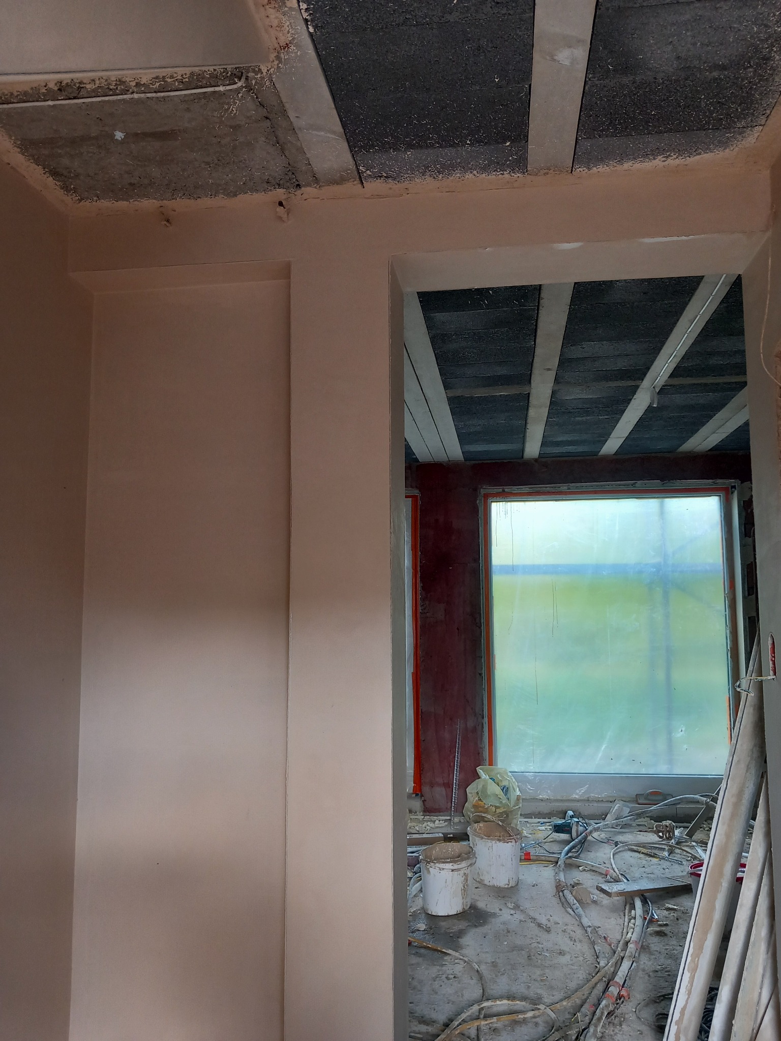 Wnętrze w trakcie prac wykończeniowych: gładkie ściany w odcieniach beżu, surowy strop z widocznymi belkami i okno z folią ochronną. Na podłodze widoczne narzędzia i materiały.