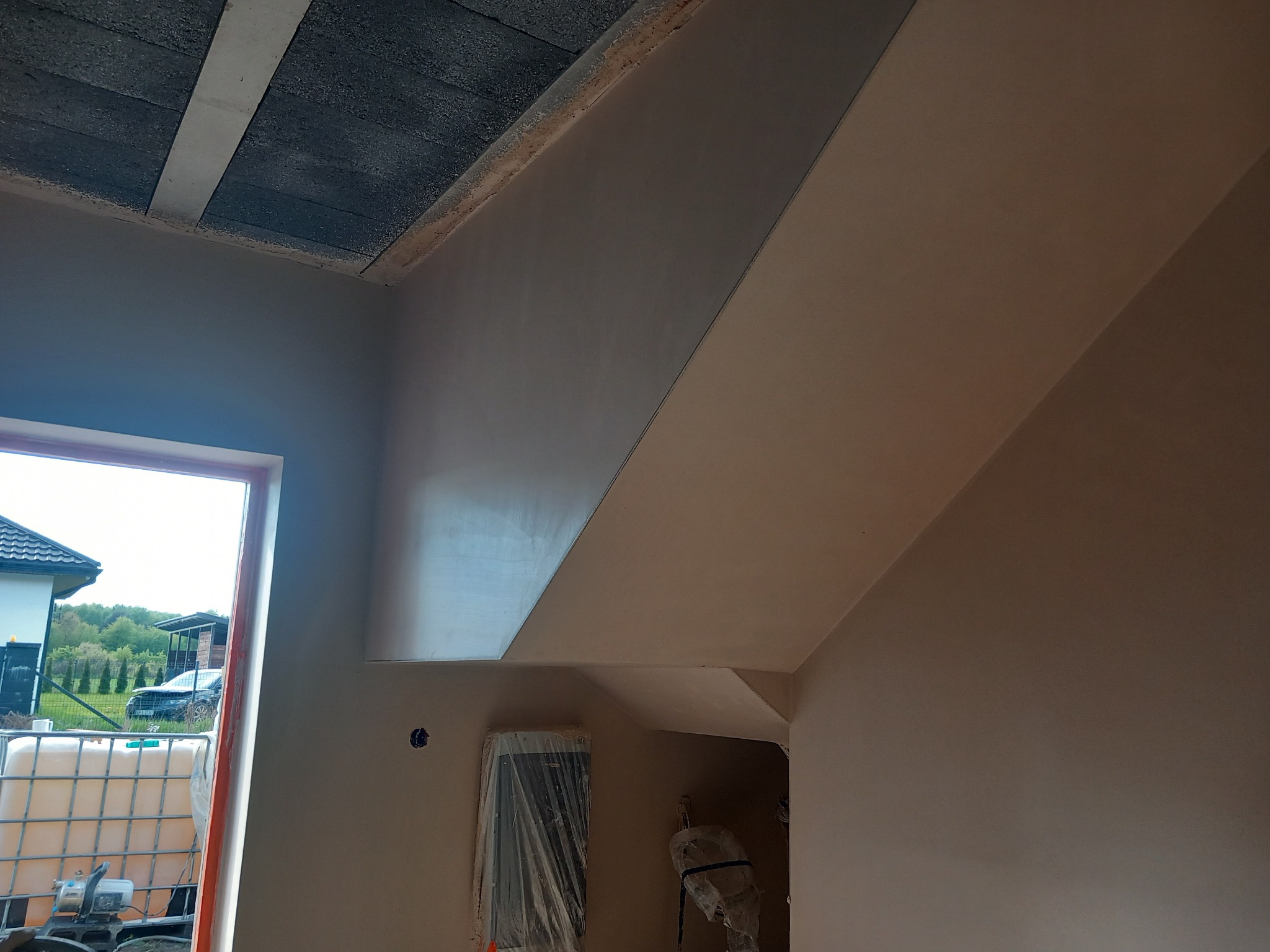 Wykończone poddasze ze ścianami pokrytymi gładzią w odcieniach beżu. Widoczne okno z widokiem na dom i ogród. Na podłodze zabezpieczone elementy instalacji.