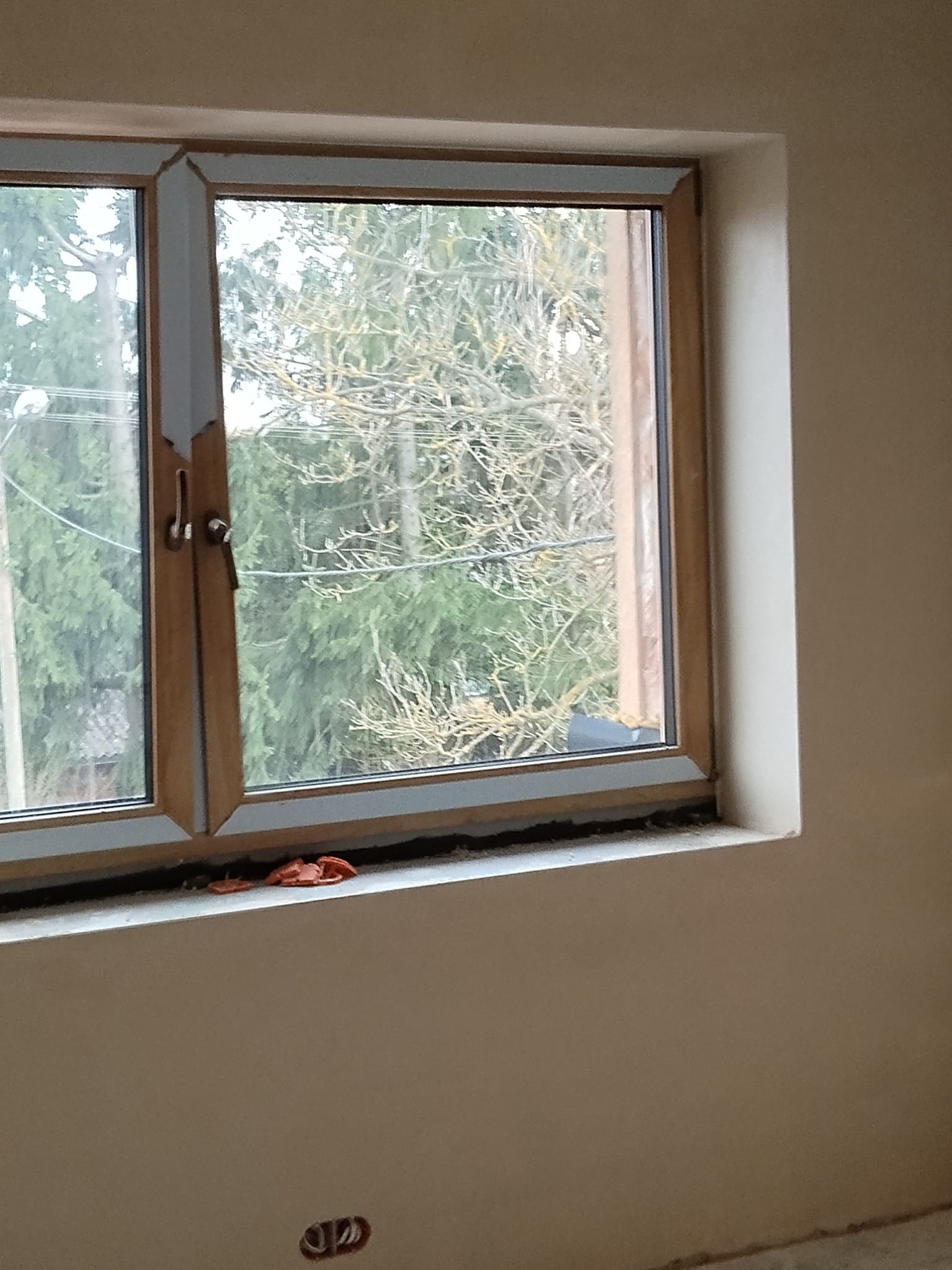 Okna z widokiem na zieleń po tynkowaniu, widoczne ślady prac na parapecie i ścianie. Nowe tynki w jasnym odcieniu, gniazdko elektryczne na dole ściany.