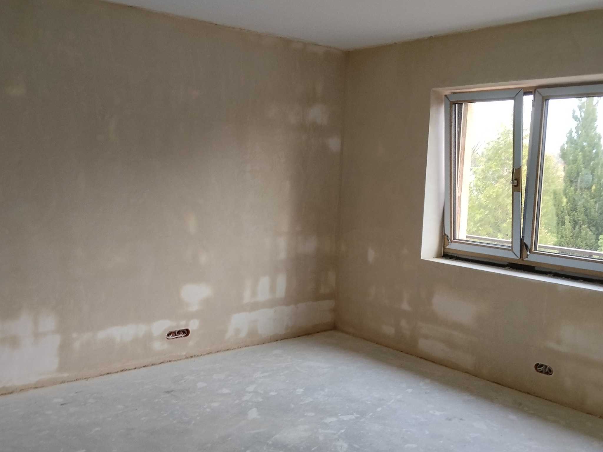 Pokój po tynkowaniu maszynowym, widoczne gładkie ściany w odcieniach beżu, okno z widokiem na zieleń i gniazdka elektryczne. Świeżo położona podłoga.