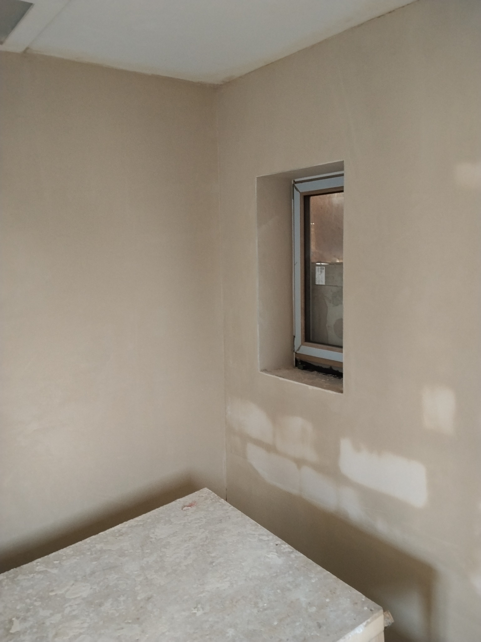 Świeżo otynkowane wnętrze z oknem, widoczne ślady po łatach. Na pierwszym planie fragment stołu roboczego. Neutralna kolorystyka ścian i sufitu.