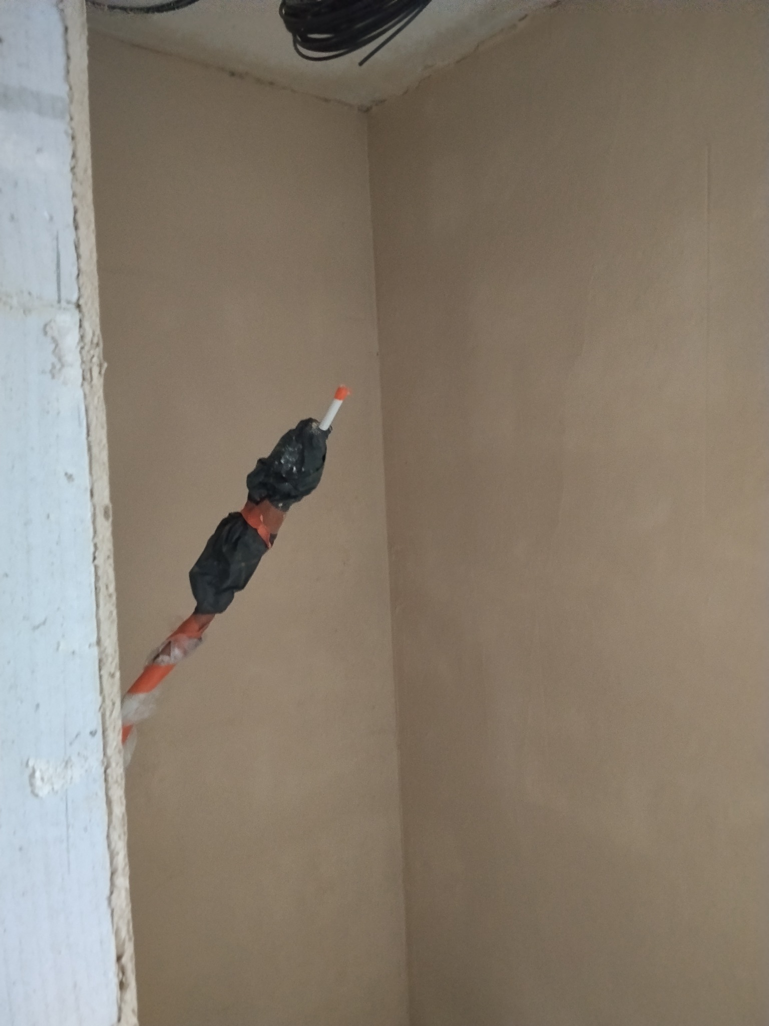 Świeżo położony tynk na ścianie w rogu pomieszczenia, widoczna wystająca instalacja elektryczna zabezpieczona taśmą. Surowy stan wykończenia wnętrza.