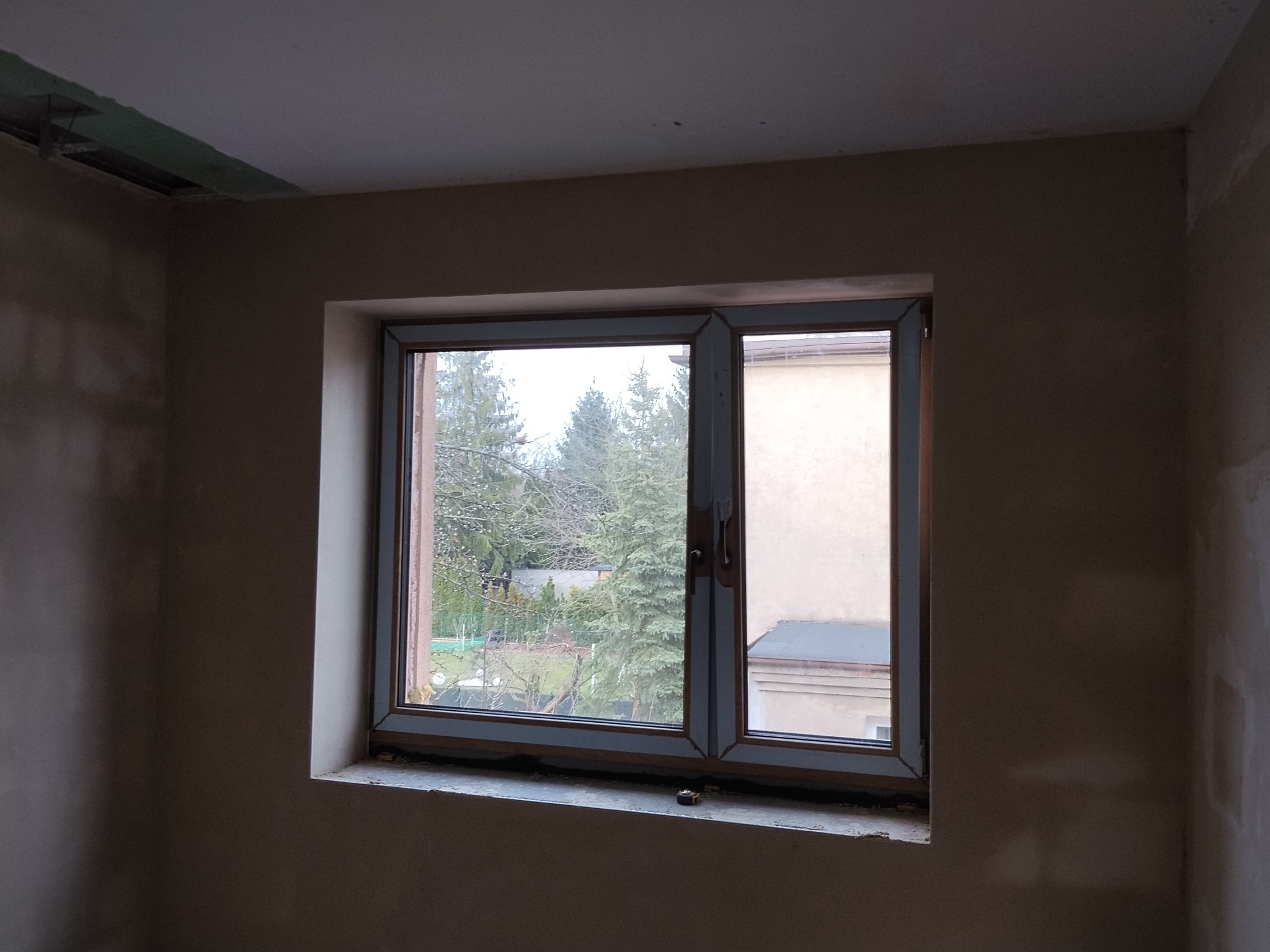 Pokój w trakcie prac wykończeniowych: ściany po nałożeniu tynku, okno z widokiem na zieleń. Widoczne niedoskonałości i surowy stan wnętrza.