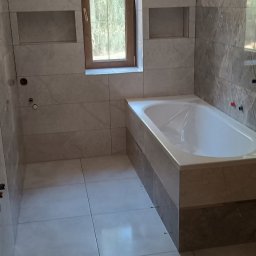 Edward Ostrołęka - Wykończona łazienka z wanną obudowaną płytkami, okno w drewnianej ramie i wnęki w ścianie. Płytki w odcieniach szarości i beżu. Widok na całe pomieszczenie.