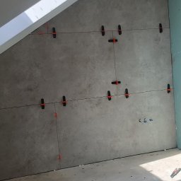 Edward Ostrołęka - Ściana w skosie z szarymi płytkami, poziomowana z użyciem klipsów i sznurka. Na podłodze wiadro z klipsami, obcęgi i miarka.