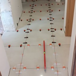 Edward Ostrołęka - Układanie jasnych płytek podłogowych z użyciem poziomicy i klipsów poziomujących w korytarzu. Widoczne narzędzia i niedokończone ściany.