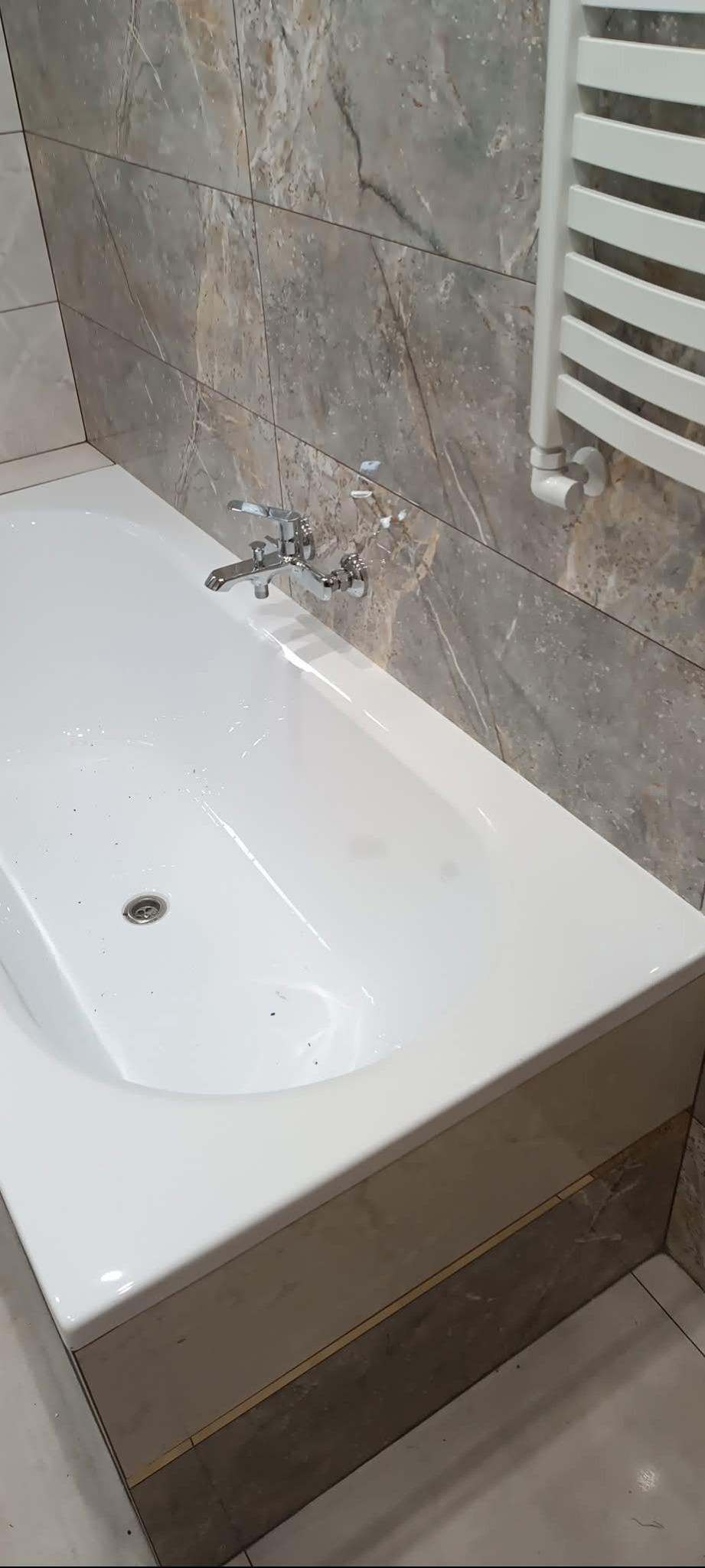 Biała wanna z chromowaną baterią na tle marmurowych płytek i grzejnika drabinkowego. Wykończenie łazienki w odcieniach szarości i bieli.