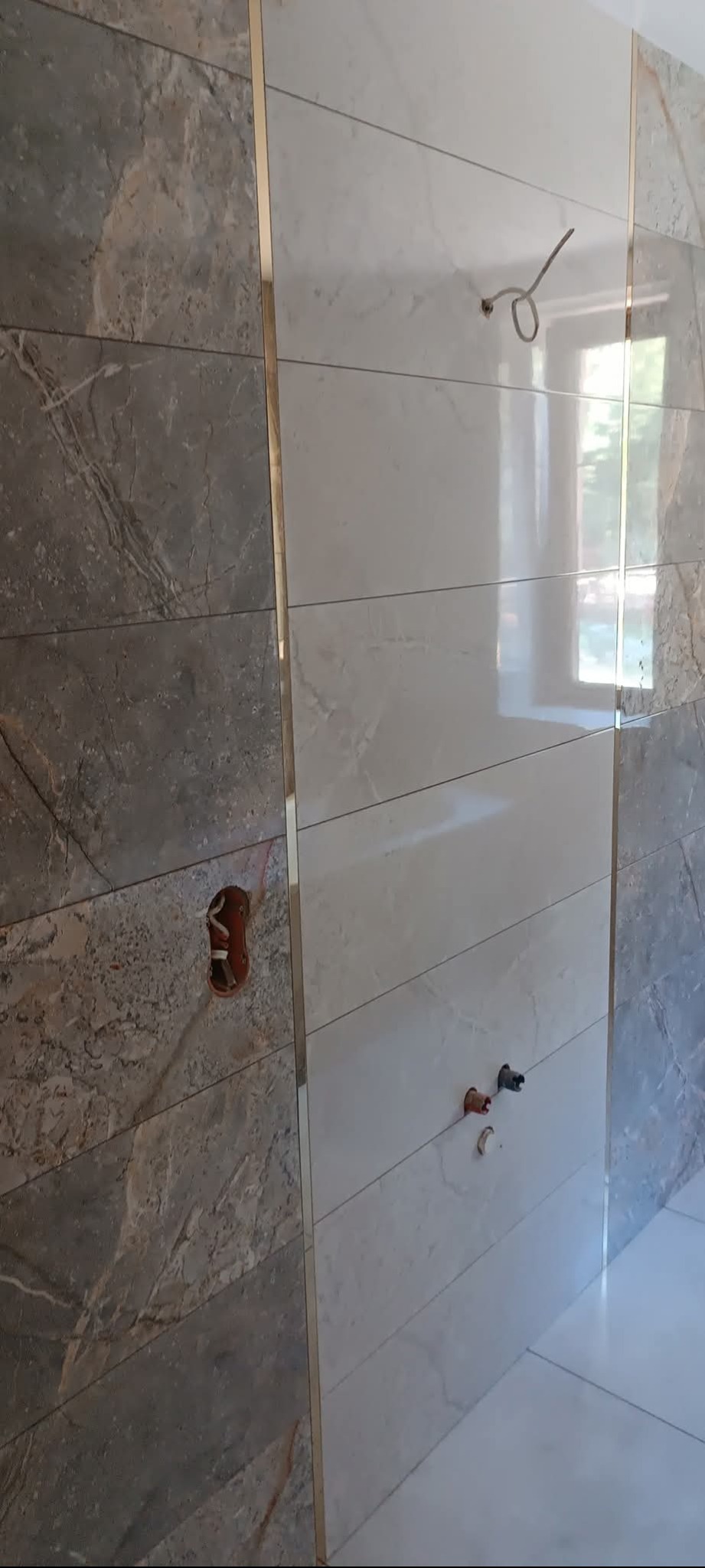 Wykończona ściana w łazience z szarych i białych płytek imitujących marmur, oddzielonych złotymi listwami. Widoczne wyprowadzenia instalacji wodnej i elektrycznej.