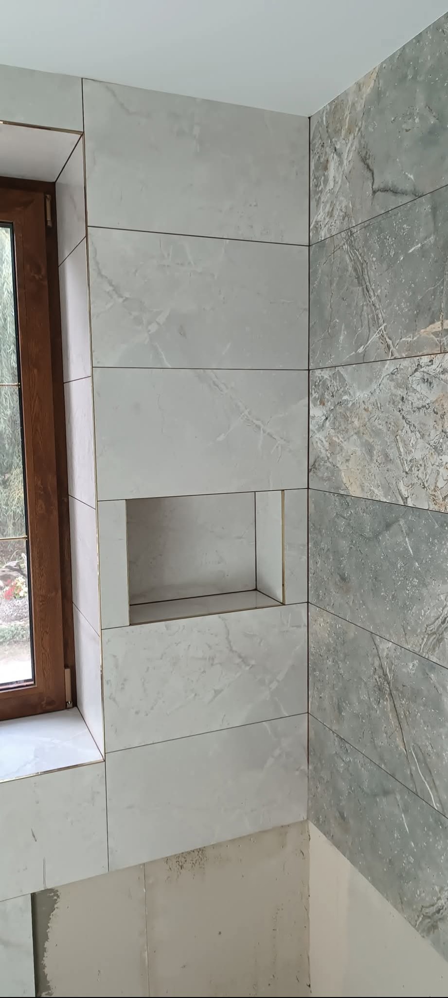 Wykończona ściana łazienki z szarych i białych płytek imitujących marmur, z wnęką obłożoną białymi płytkami, widoczny fragment okna.