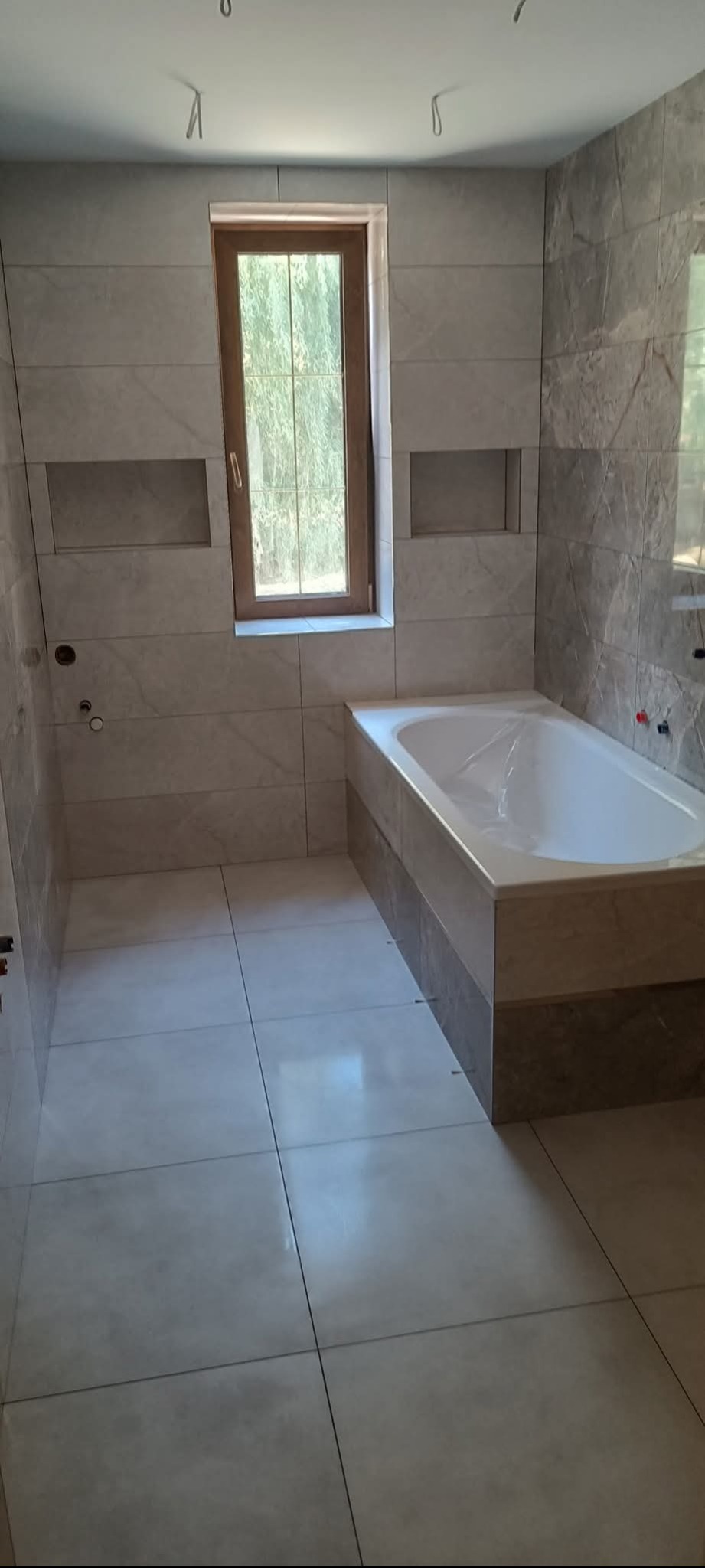 Wykończona łazienka z wanną obudowaną płytkami, okno w drewnianej ramie i wnęki w ścianie. Płytki w odcieniach szarości i beżu. Widok na całe pomieszczenie.