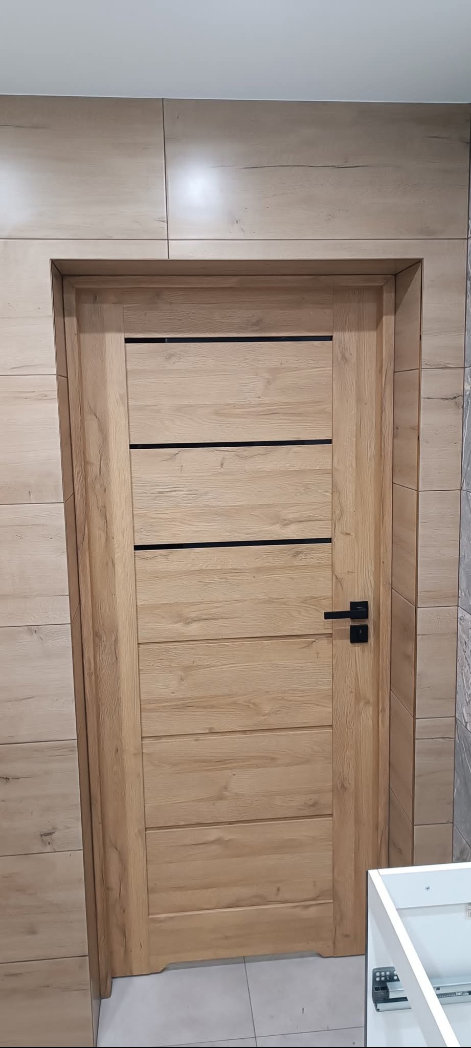 Zamknięte, drewniane drzwi wewnętrzne z czarną klamką, wkomponowane w ścianę wyłożoną płytkami imitującymi drewno. Widoczny fragment białej szafki.