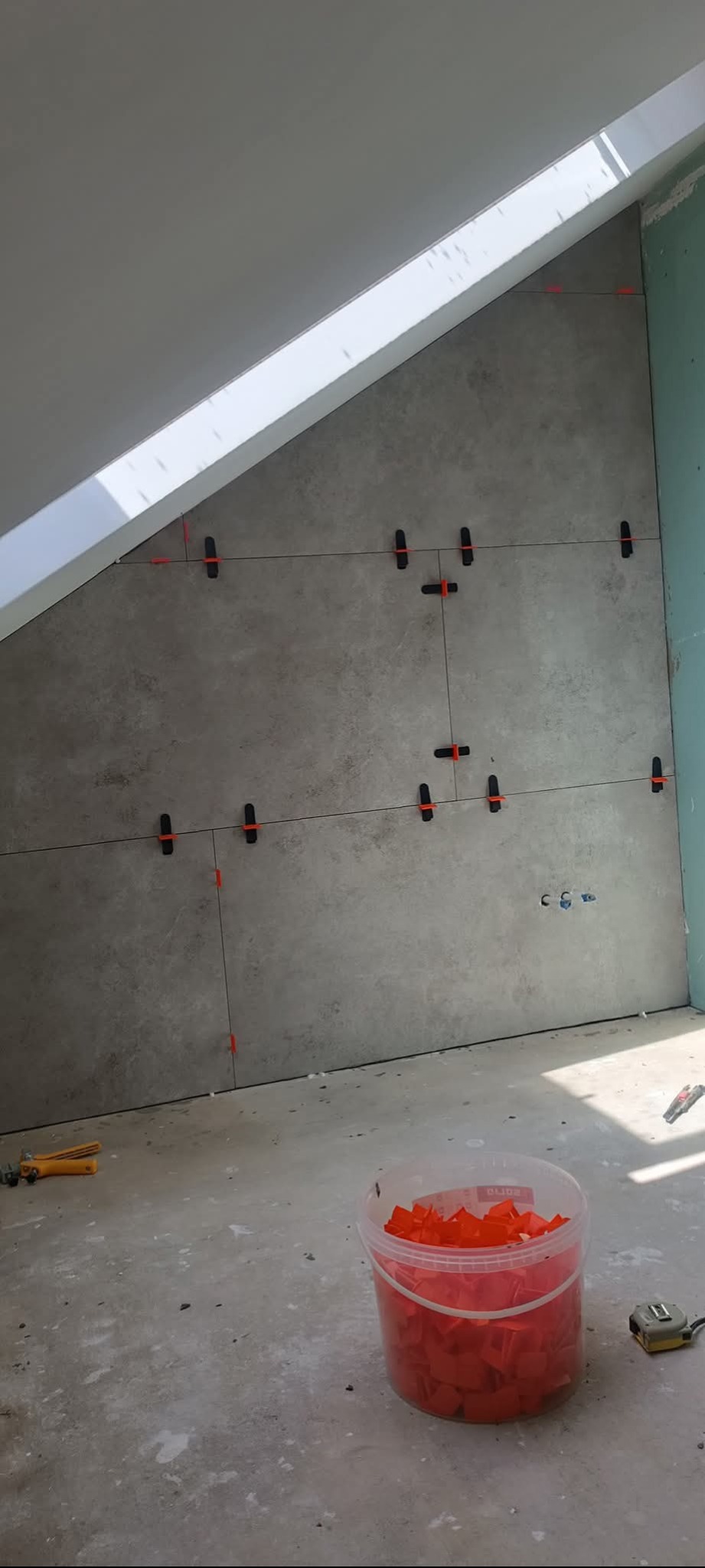 Ściana w skosie z szarymi płytkami, poziomowana z użyciem klipsów i sznurka. Na podłodze wiadro z klipsami, obcęgi i miarka.