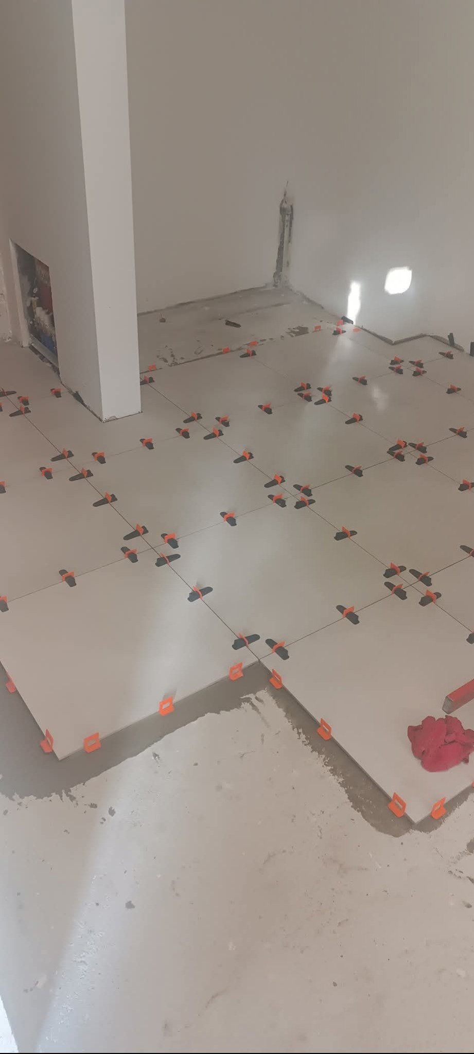 Układanie jasnych płytek podłogowych z użyciem pomarańczowych klipsów poziomujących w trakcie remontu. Widoczne fugi i narzędzia glazurnicze.