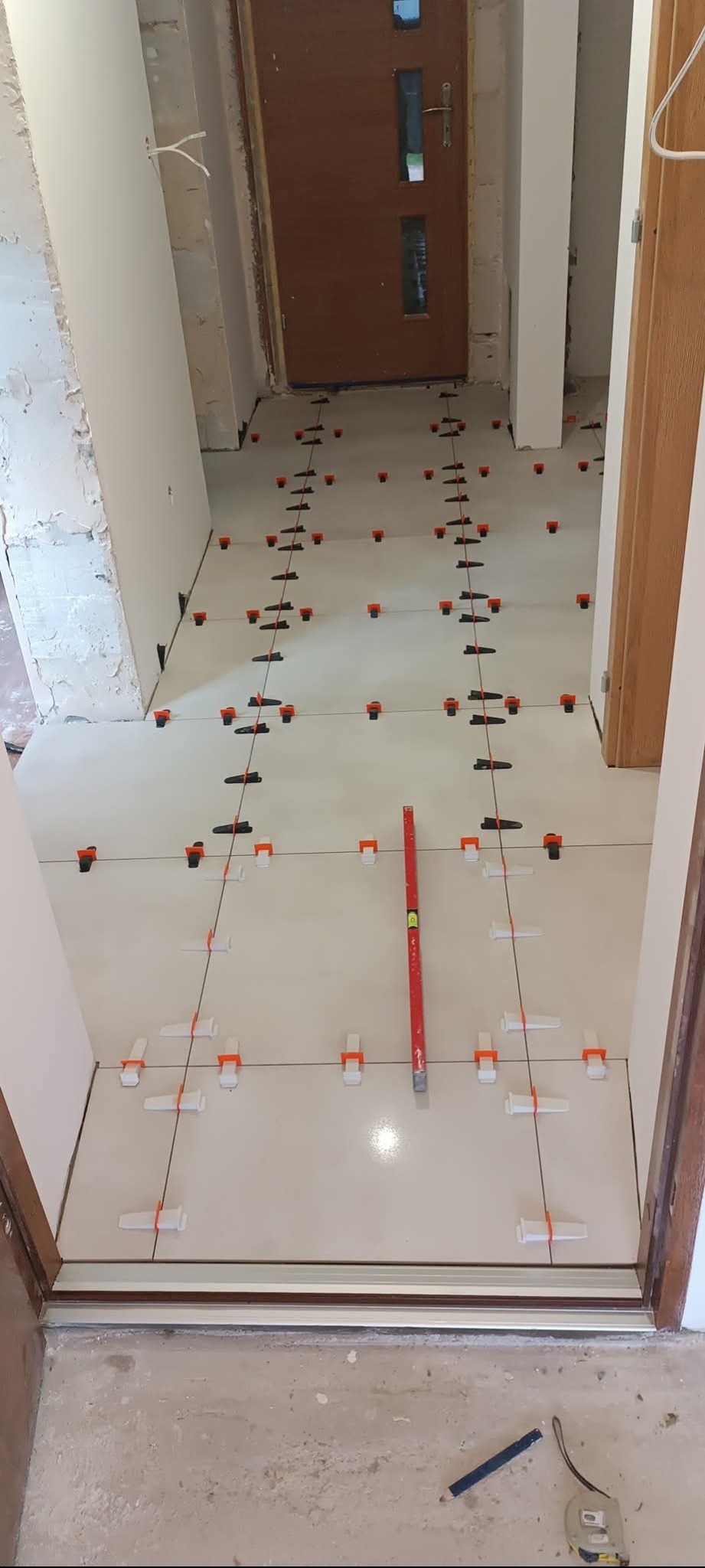 Układanie jasnych płytek podłogowych z użyciem poziomicy i klipsów poziomujących w korytarzu. Widoczne narzędzia i niedokończone ściany.