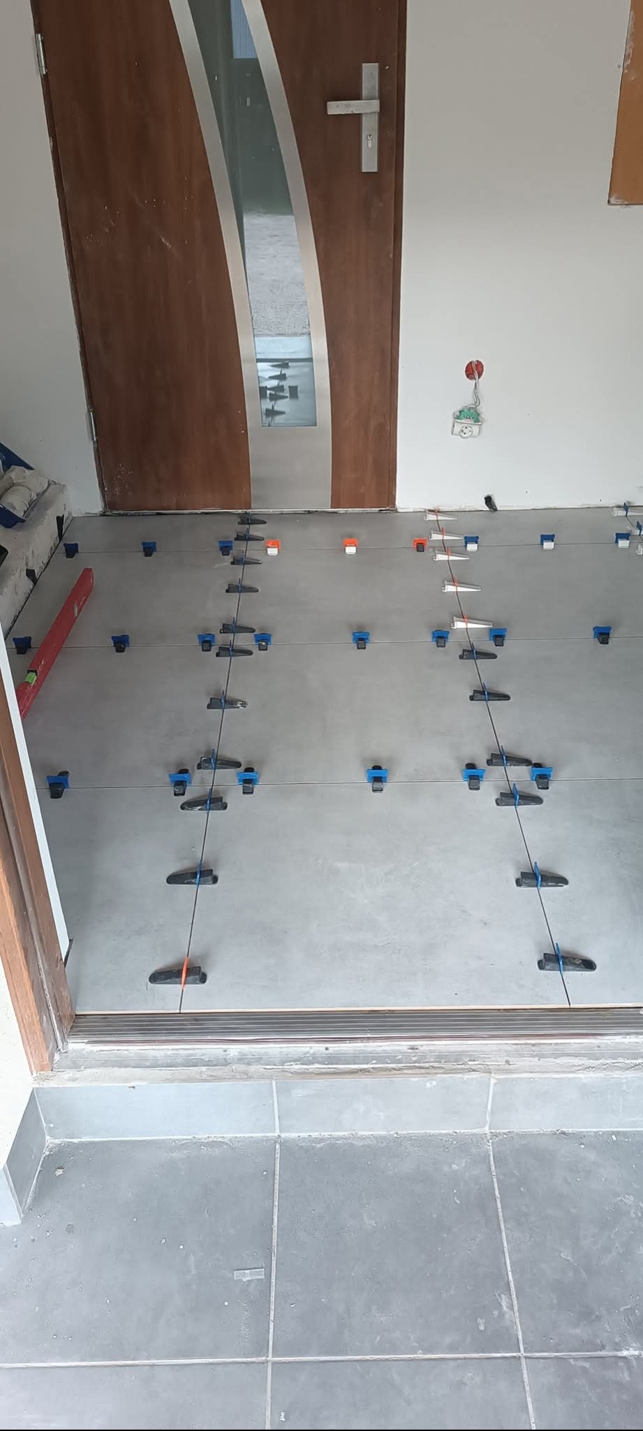 Układanie szarych płytek podłogowych z użyciem systemu poziomowania. Widoczna poziomica i drzwi z przeszkleniem.