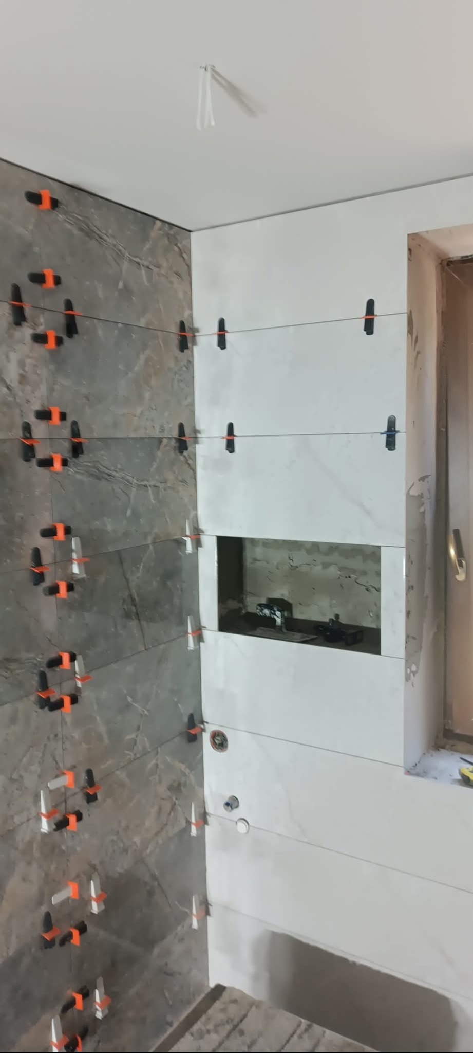 Układanie płytek w łazience z użyciem systemu poziomowania. Widoczne szare i białe płytki, sznurki i klipsy. Fragment okna w tle.