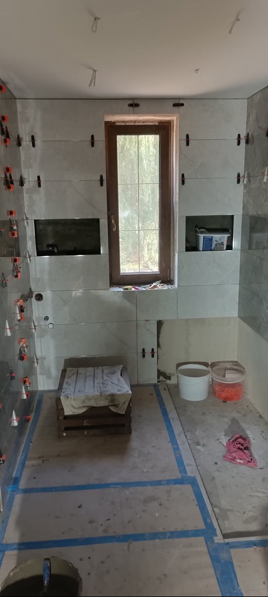 Łazienka w trakcie remontu: jasne płytki na ścianach z systemem poziomowania, okno z drewnianą ramą, przygotowane wnęki. Podłoga z wyznaczonym obszarem do dalszych prac.