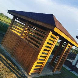 Świat Drewna - Drewniana altana z brązowym dachem, oświetlona popołudniowym słońcem, z widocznymi detalami konstrukcyjnymi i otoczeniem zieleni.