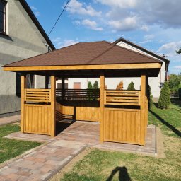 Świat Drewna - Drewniana altana z brązowym dachem na trawniku, obok domu. Konstrukcja z jasnego drewna, częściowo zabudowana. Ścieżka z kostki prowadzi do wejścia.