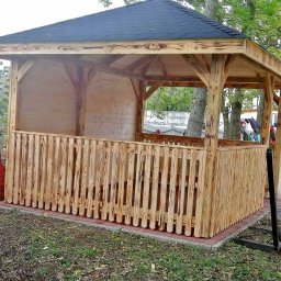 Świat Drewna - Drewniana altana z czarnym dachem, otoczona zielenią. Częściowo zabudowana, z niskim płotkiem z pionowych desek. Widoczne słoje drewna i ceglana podstawa.