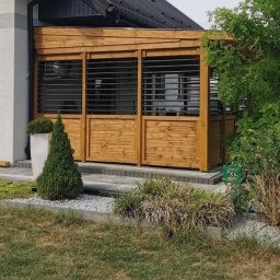 Świat Drewna - Drewniana zabudowa tarasu z żaluzjami w nowoczesnym stylu, przylegająca do domu. Uwagę zwraca jasne drewno i minimalistyczny design, wkomponowany w otoczenie ogrodu.