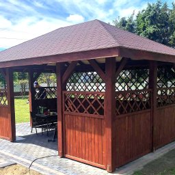 Świat Drewna - Drewniana altana z brązowym dachem i ażurowymi ściankami, ustawiona na kostce brukowej w ogrodzie. Widoczne meble ogrodowe wewnątrz.