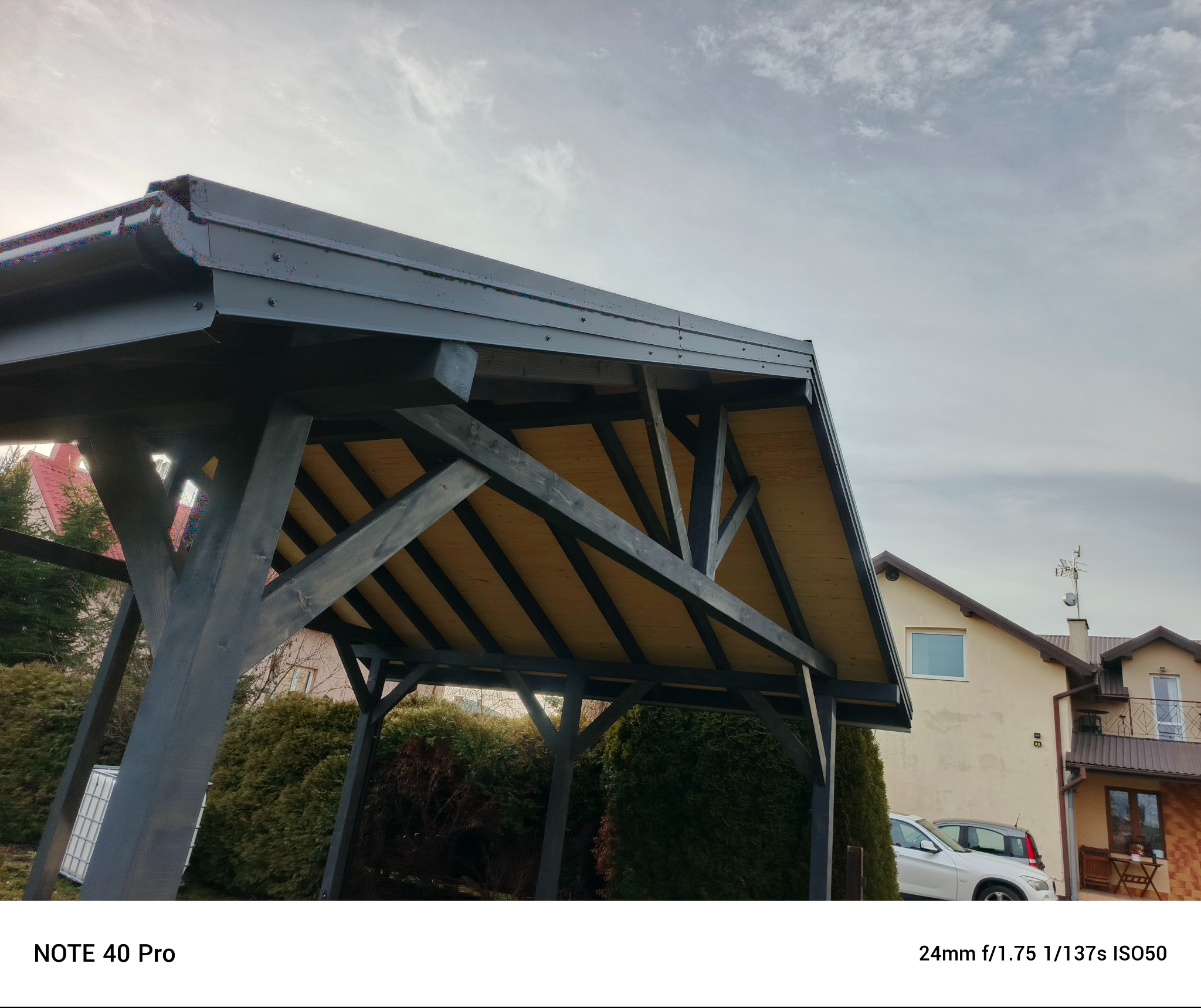 Czarna, drewniana pergola z widoczną konstrukcją dachu i elementami mocującymi. W tle dom i zieleń. Ujęcie z dołu, podkreślające solidność konstrukcji.