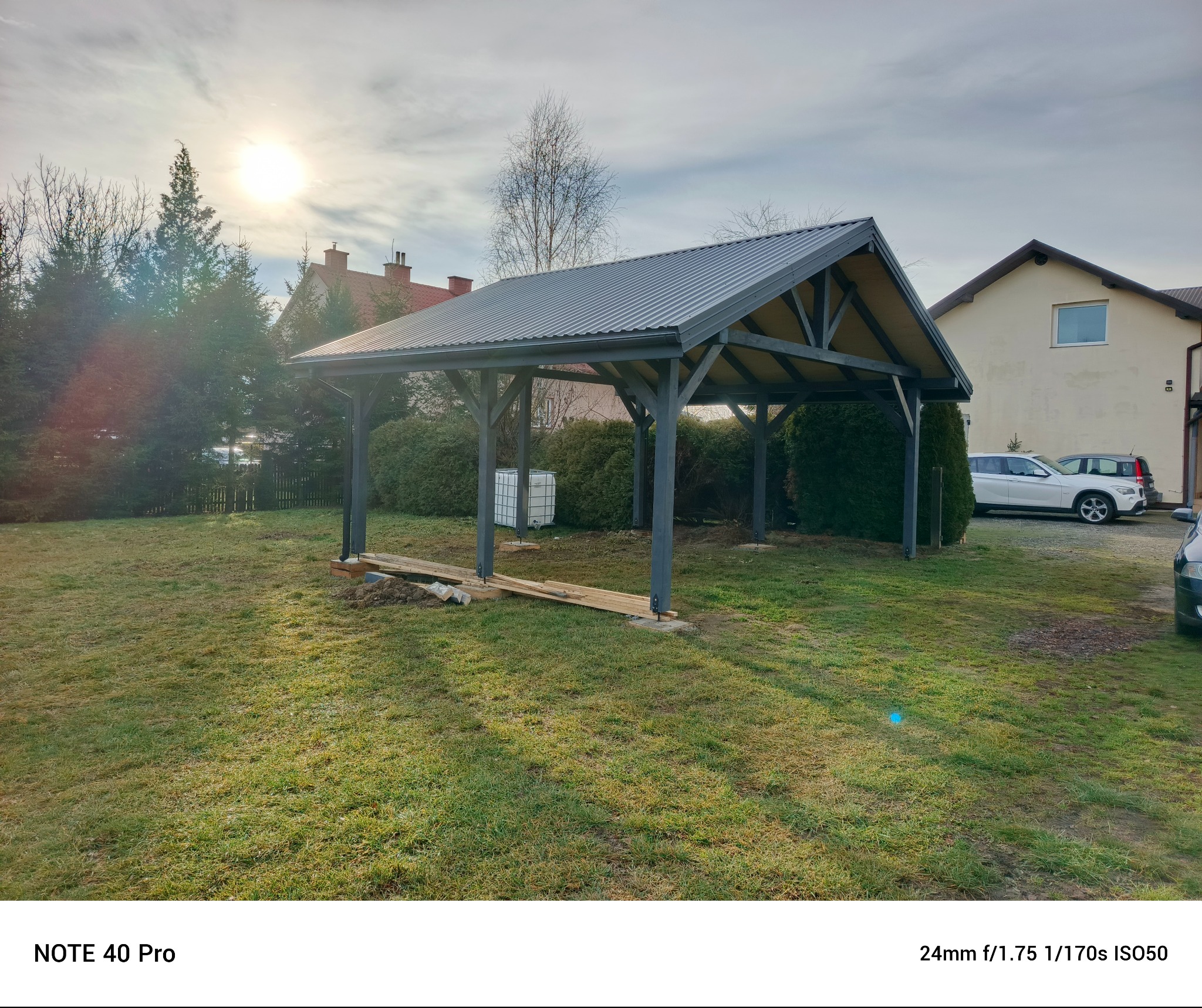 Szara, metalowa altana z drewnianą konstrukcją w ogrodzie, w trakcie montażu. Widoczne deski i fundamenty. W tle dom i zieleń.