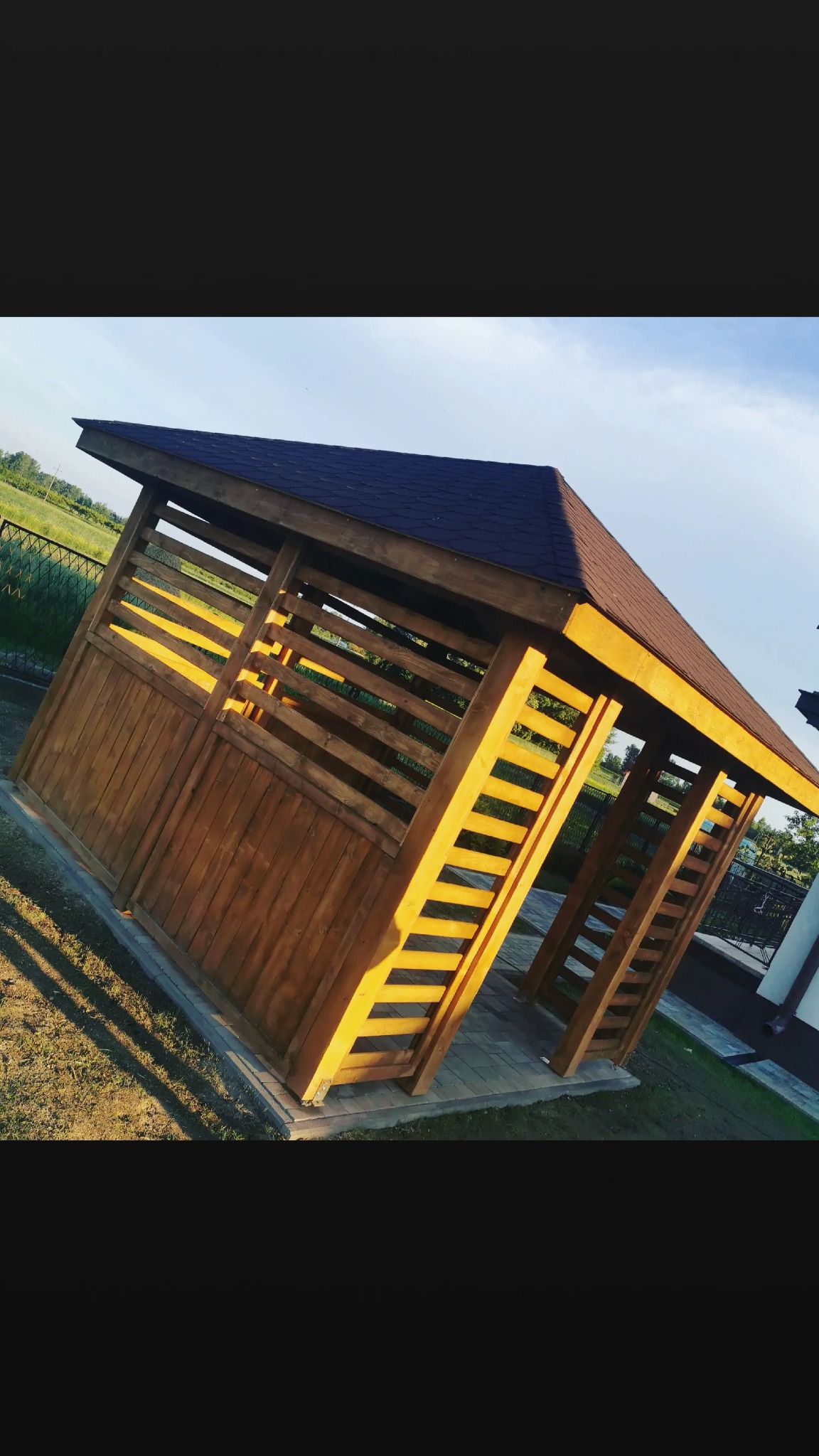 Drewniana altana z brązowym dachem, oświetlona popołudniowym słońcem, z widocznymi detalami konstrukcyjnymi i otoczeniem zieleni.