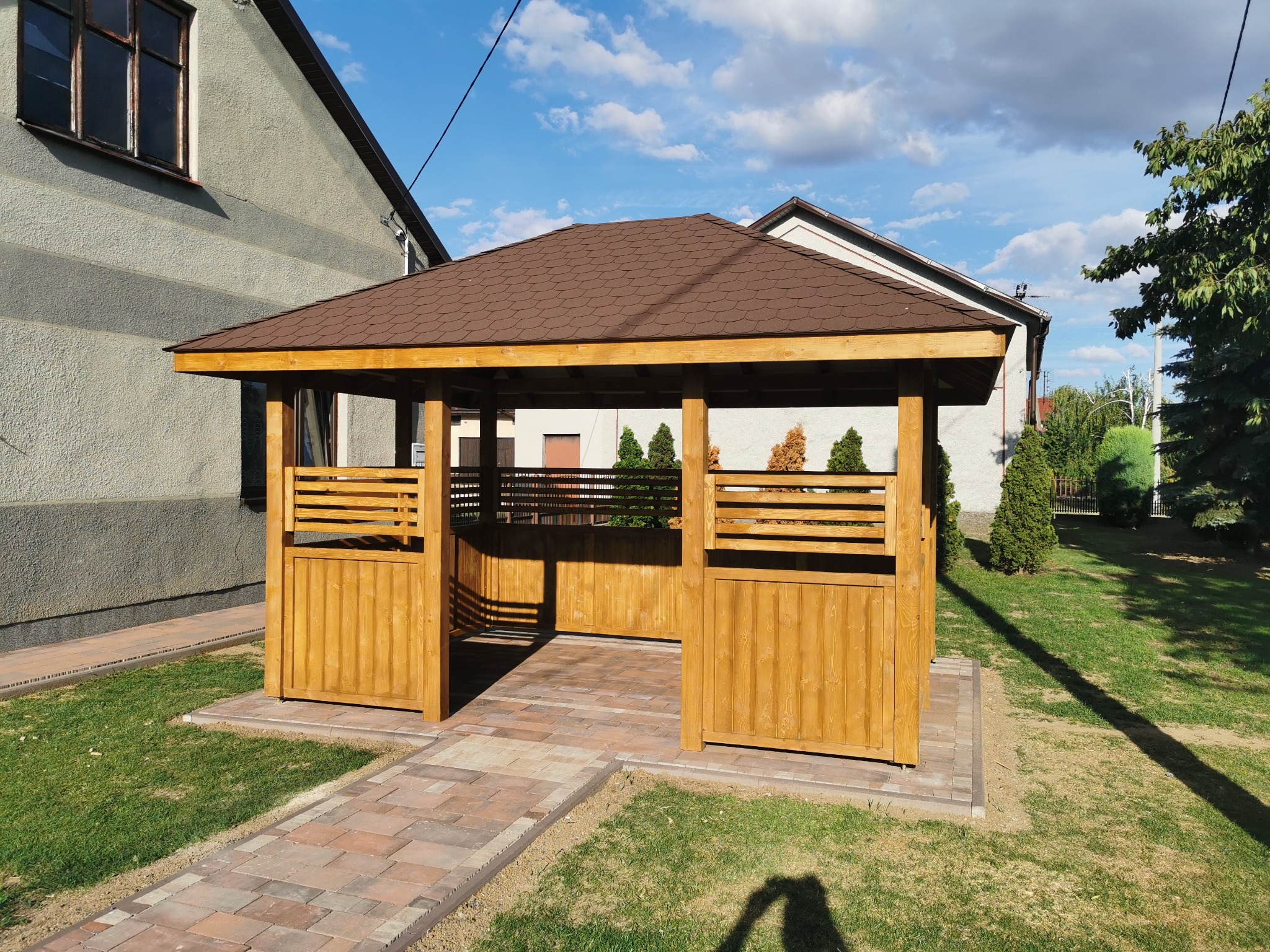 Drewniana altana z brązowym dachem na trawniku, obok domu. Konstrukcja z jasnego drewna, częściowo zabudowana. Ścieżka z kostki prowadzi do wejścia.