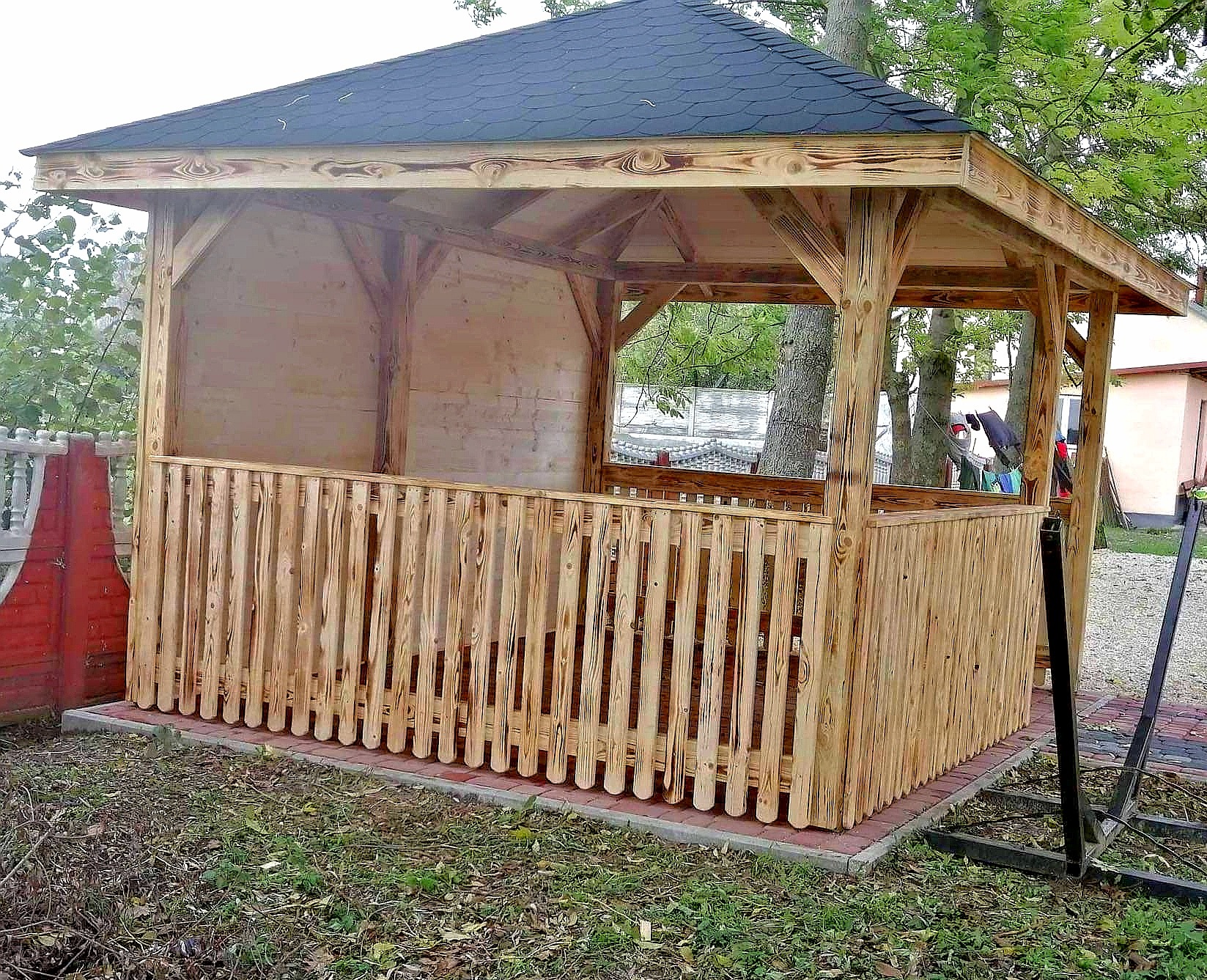 Drewniana altana z czarnym dachem, otoczona zielenią. Częściowo zabudowana, z niskim płotkiem z pionowych desek. Widoczne słoje drewna i ceglana podstawa.