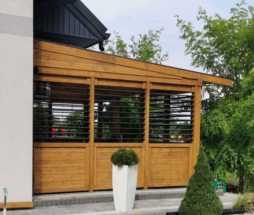 Drewniana altana z żaluzjami w ogrodzie, widok z zewnątrz. Jasne drewno, nowoczesny design, rośliny w donicach. Elementy czarnego dachu widoczne w tle.