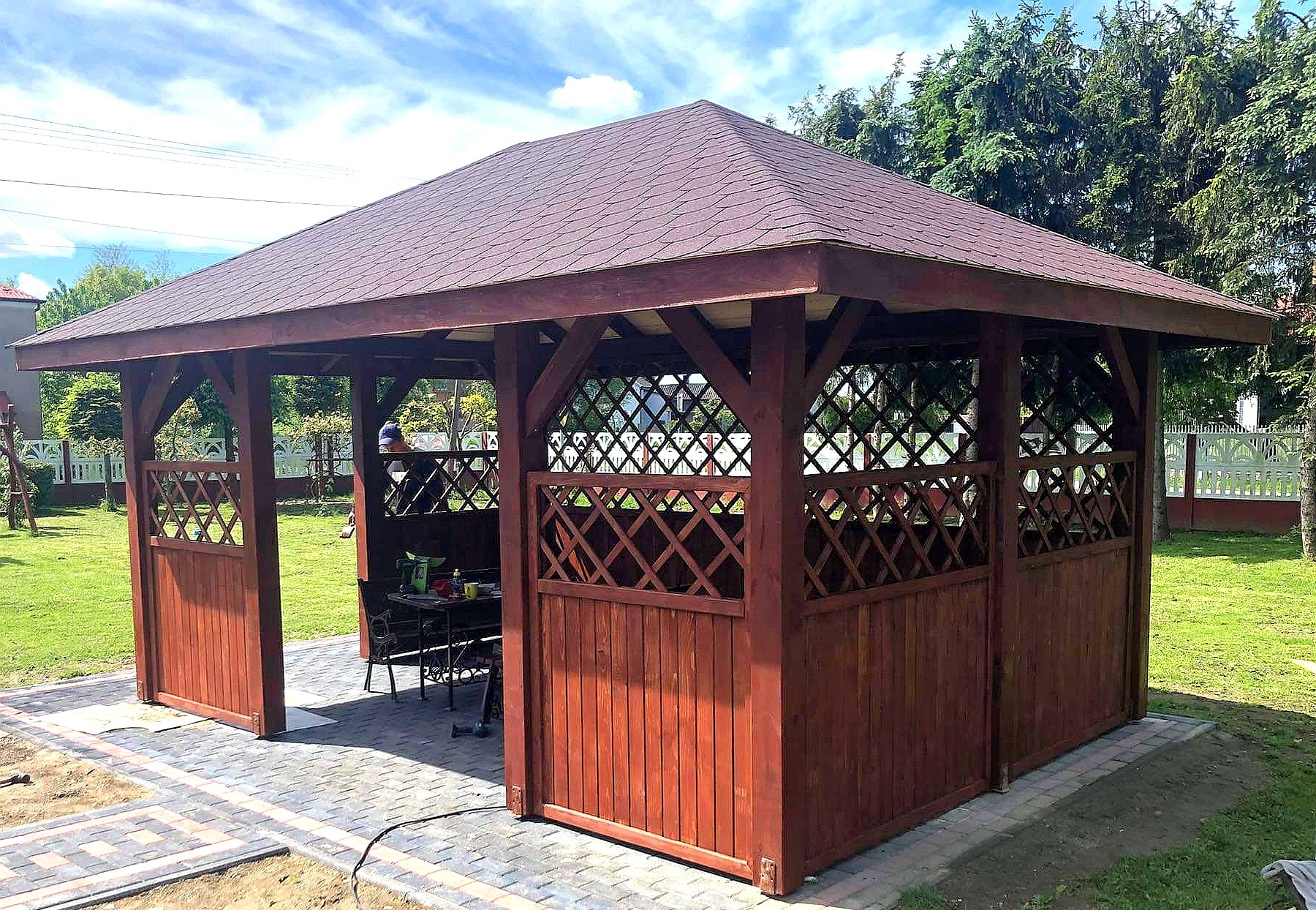 Drewniana altana z brązowym dachem i ażurowymi ściankami, ustawiona na kostce brukowej w ogrodzie. Widoczne meble ogrodowe wewnątrz.
