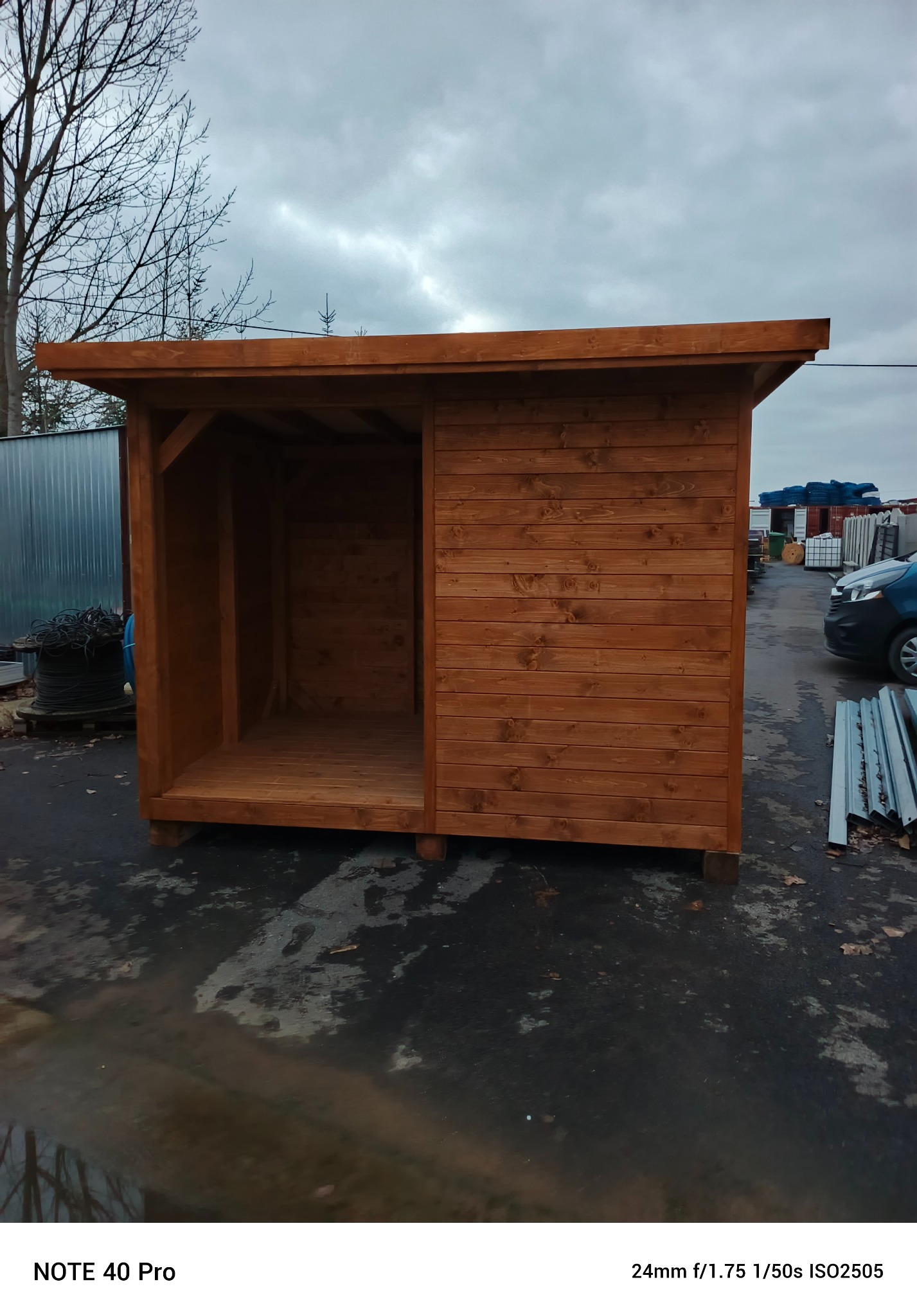 Drewniana konstrukcja z brązowych desek, przypominająca małą altanę lub schowek, z otwartym wejściem z jednej strony, na zewnątrz, w pochmurny dzień.