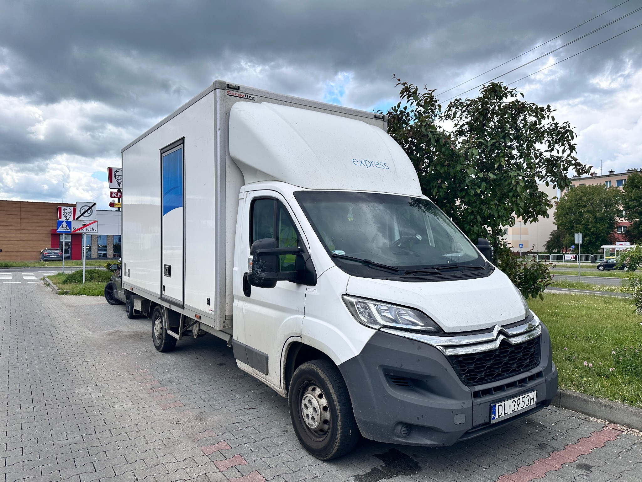 Biały samochód dostawczy z nadrukiem 'express' zaparkowany na chodniku, widoczny budynek KFC w tle. Rejestracja DL 3953H.