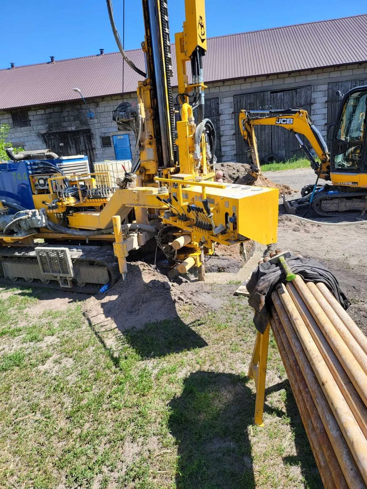 Żółta wiertnica w trakcie pracy, z widocznymi rurami wiertniczymi i koparką JCB w tle, na tle budynku z cegły i błękitnego nieba.