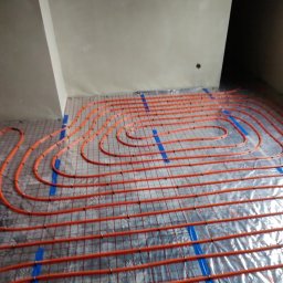INSTAL-KAM Karol Kamiński - Pomarańczowe rury ogrzewania podłogowego ułożone na folii izolacyjnej, przymocowane niebieską taśmą do siatki. Widok z góry na instalację w surowym wnętrzu.
