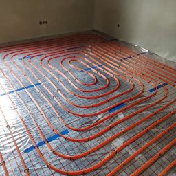 INSTAL-KAM Karol Kamiński - Pomarańczowe rury ogrzewania podłogowego ułożone na srebrnej folii z czerwoną siatką, przymocowane niebieską taśmą, widok z góry na surową ścianę i poddasze.