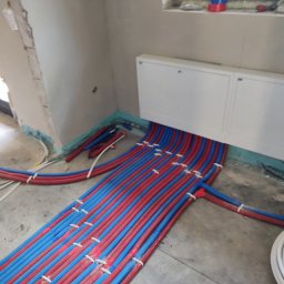 INSTAL-KAM Karol Kamiński - Instalacja ogrzewania podłogowego z rurami w kolorze czerwonym i niebieskim, ułożonymi równolegle na szarym betonie, przed wylaniem posadzki. Widoczne białe szafki.