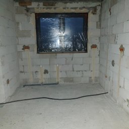 INSTAL-KAM Karol Kamiński - Surowy stan: ściany z betonu komórkowego z instalacją elektryczną i rurami, okno zabezpieczone folią. W tle widoczne przewody na podłodze.