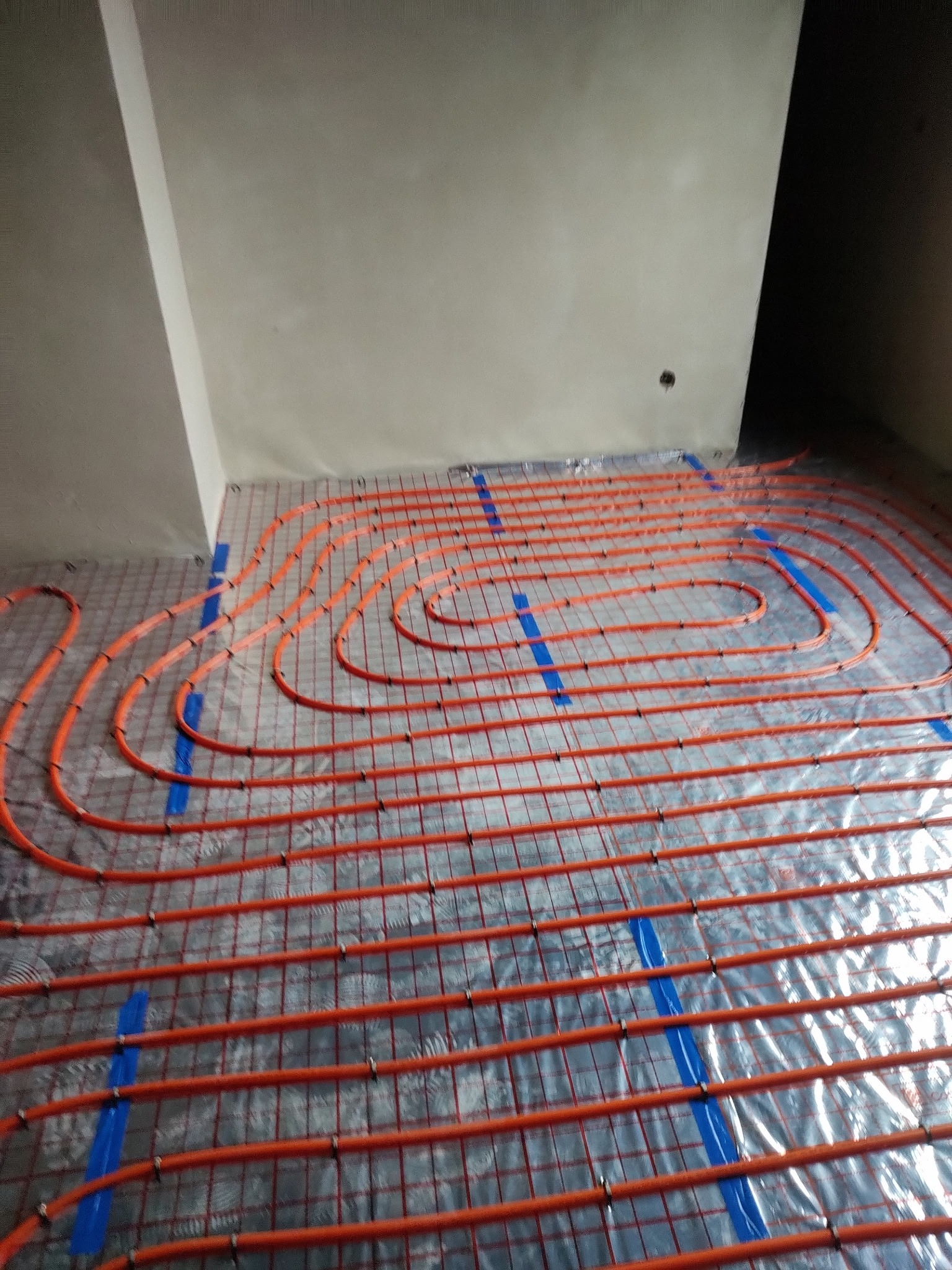 Pomarańczowe rury ogrzewania podłogowego ułożone na folii izolacyjnej, przymocowane niebieską taśmą do siatki. Widok z góry na instalację w surowym wnętrzu.
