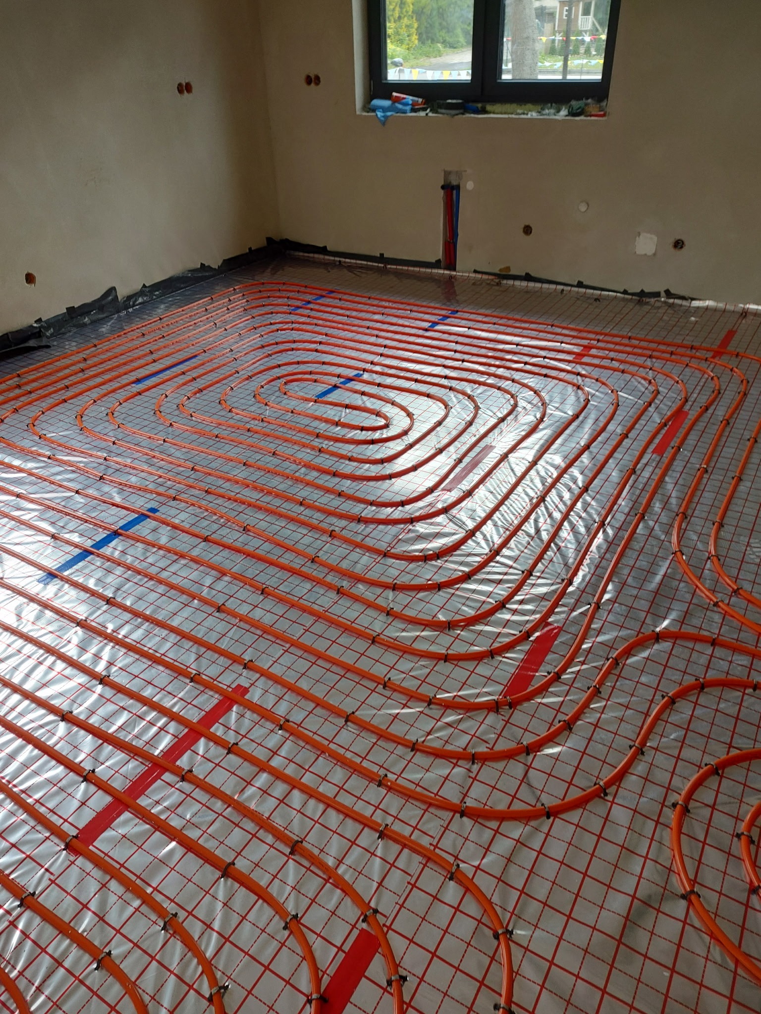 Pomarańczowe rury ogrzewania podłogowego ułożone spiralnie na folii z siatką w jasnym pomieszczeniu z oknem. Widoczne mocowania rur i fragmenty ścian.