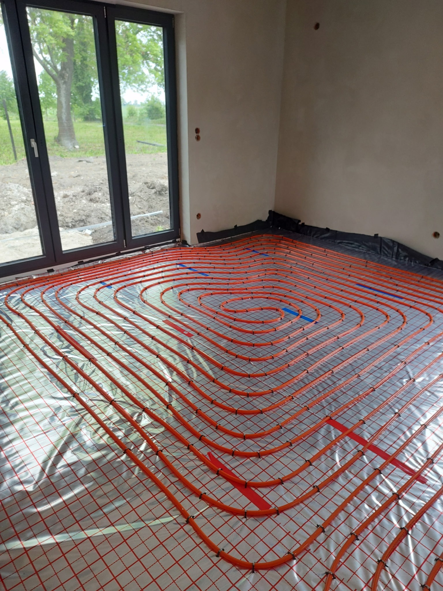 Pomarańczowe rury ogrzewania podłogowego ułożone spiralnie na srebrnej folii w jasnym pomieszczeniu z dużym oknem i widokiem na zieleń na zewnątrz.